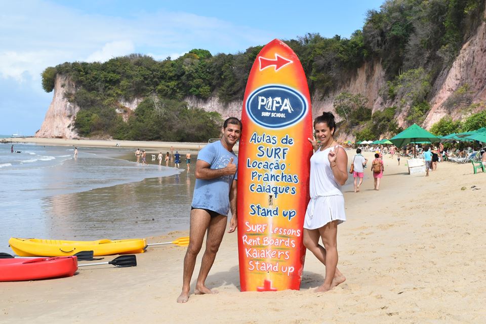 Visitantes ao lado da prancha com a identificação da escola na praia
